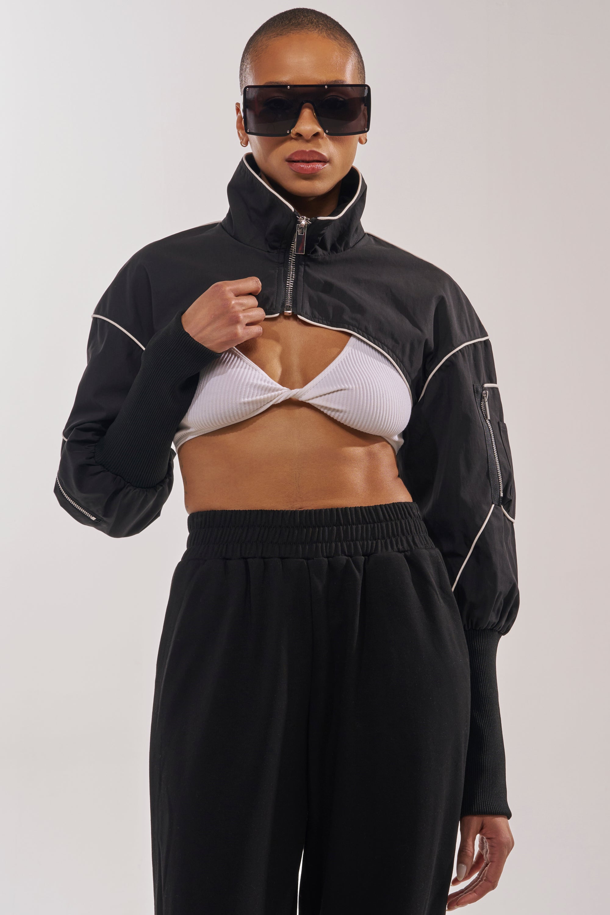 A person displays street style layering with oversized sunglasses, the NOW YOU SEE ME SPORTY BOLERO over a white ribbed bralette, and black high-waisted pants against a plain background. 1