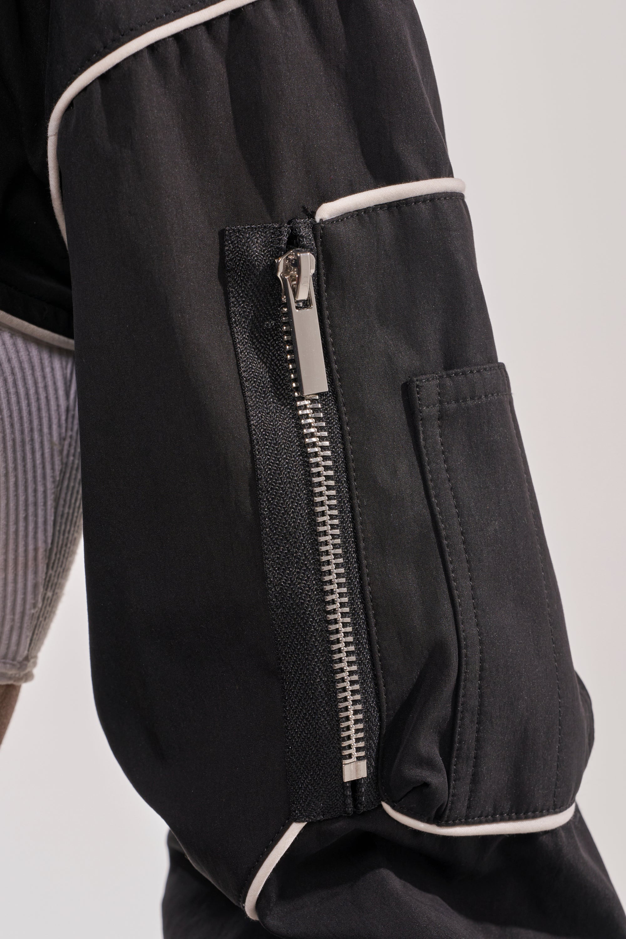 Close-up of NOW YOU SEE ME SPORTY BOLERO’s black sleeve with white piping, a vertical silver zipper, and a small rectangular side pocket. The background is plain and light-colored. 9