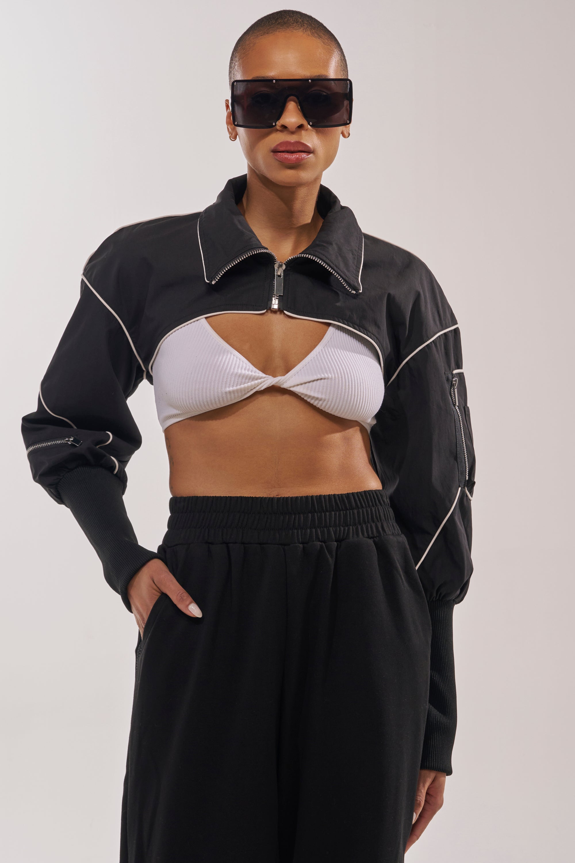 Wearing black sunglasses, the person stands confidently with one hand in their pocket, styled in the NOW YOU SEE ME SPORTY BOLERO— a black, zip-front jacket with white trim—paired with a white ribbed bralette and black high-waisted pants. 2