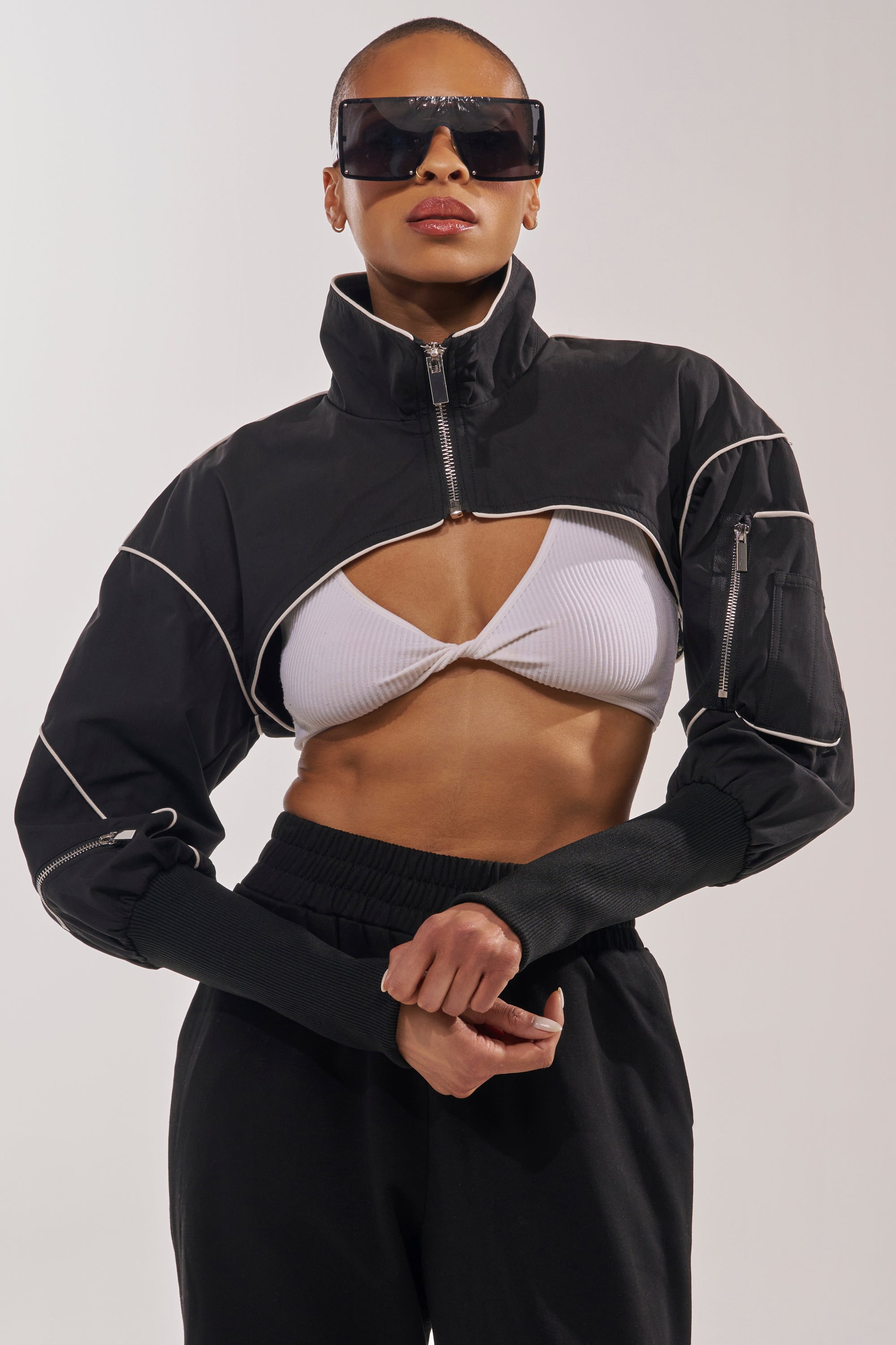 A woman stands with arms crossed in front of a neutral background, wearing large black sunglasses, the NOW YOU SEE ME SPORTY BOLERO with white piping, a white ribbed cutout crop top, and black pants. 4