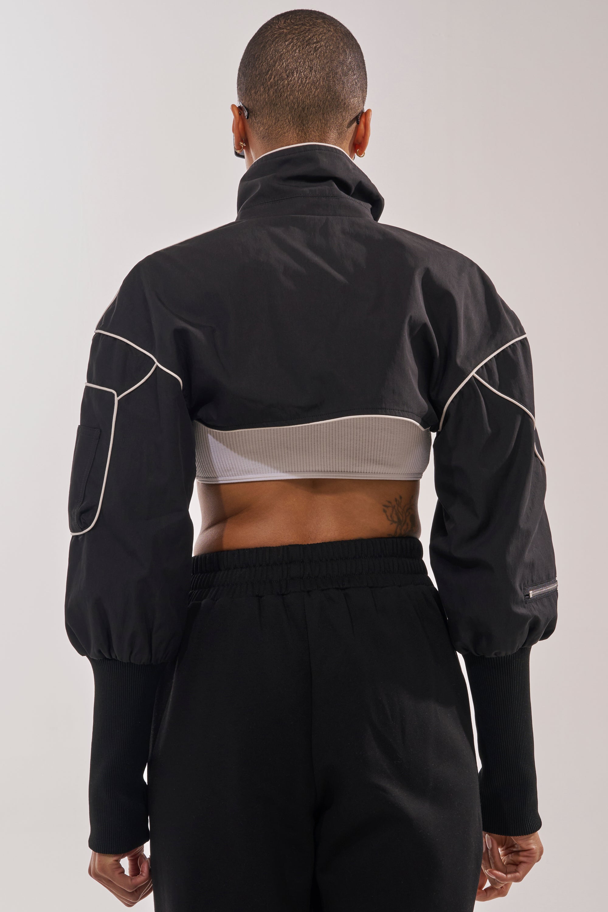 A person with closely shaved hair faces away, wearing the NOW YOU SEE ME SPORTY BOLERO—a black zip-front jacket with white piping and puffy sleeves—over a gray ribbed crop top, loose black pants, and showing a lower back tattoo. 6