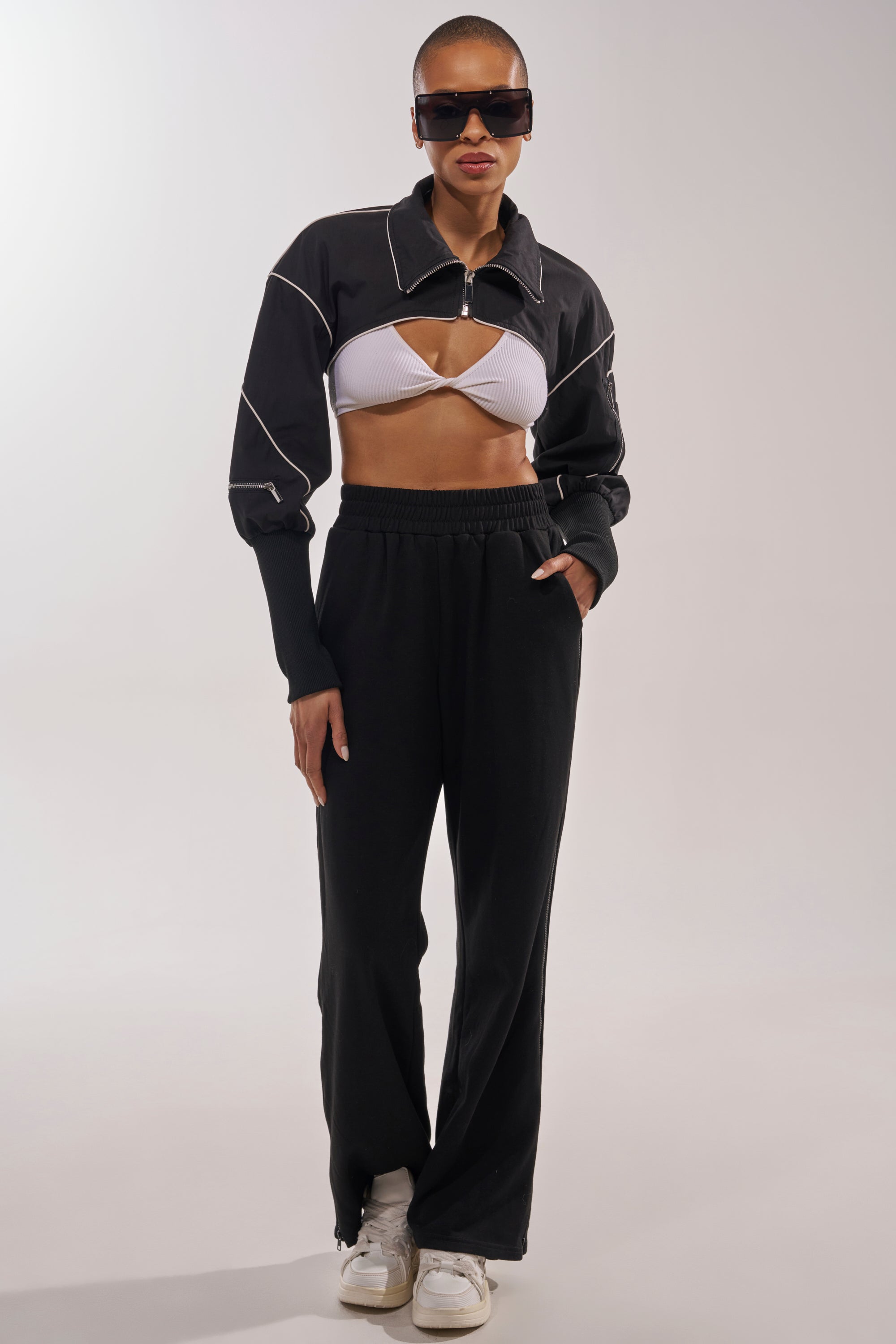 A woman stands confidently with one hand in her pocket, wearing the NOW YOU SEE ME SPORTY BOLERO over a white sports bra, paired with black sunglasses, black high-waisted pants, and white sneakers against a plain background. 7