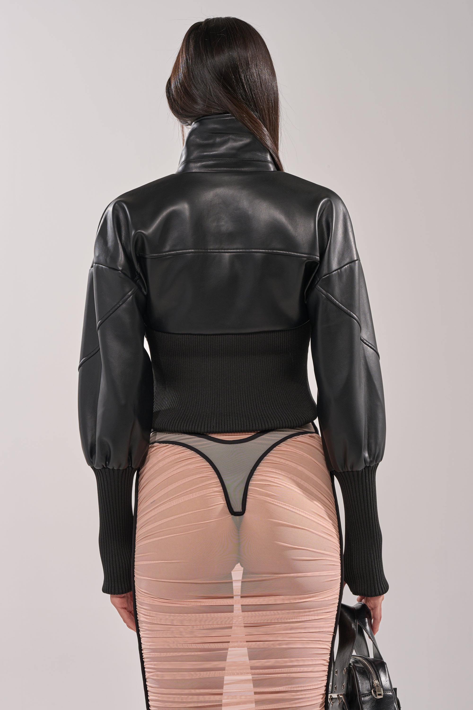 A person with long brown hair faces away, wearing the SAB FITTED MOTO jacket with ribbed sleeves and a sheer nude ruched skirt over a black thong, holding a black handbag in their right hand.