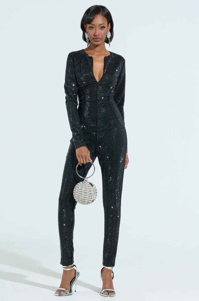 Wearing a SPARKLE SPARKLE RHINESTONE LONG SLEEVE JUMPSUIT and high heels, a confident person poses with a figure-flattering fit. They hold a round silver beaded clutch and wear statement earrings against a plain, light-colored background. 1