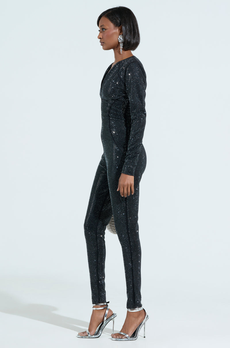 A person with shoulder-length hair stands in profile against a plain background, wearing a SPARKLE SPARKLE RHINESTONE LONG SLEEVE JUMPSUIT and silver high heels. Large earrings add to the look's elegance. 2