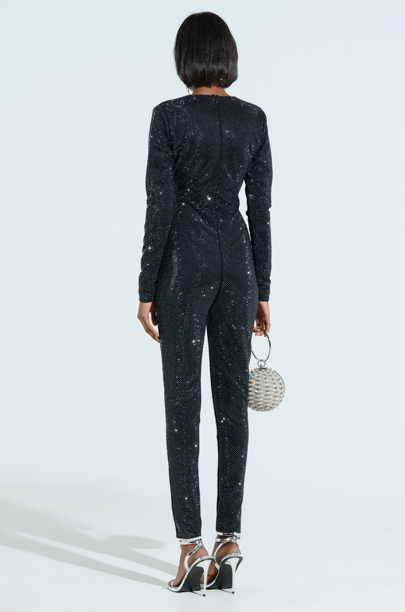 Facing away from the camera, a person showcases the SPARKLE SPARKLE RHINESTONE LONG SLEEVE JUMPSUIT paired with a round, textured handbag and silver high-heeled sandals against a plain, light background. 3