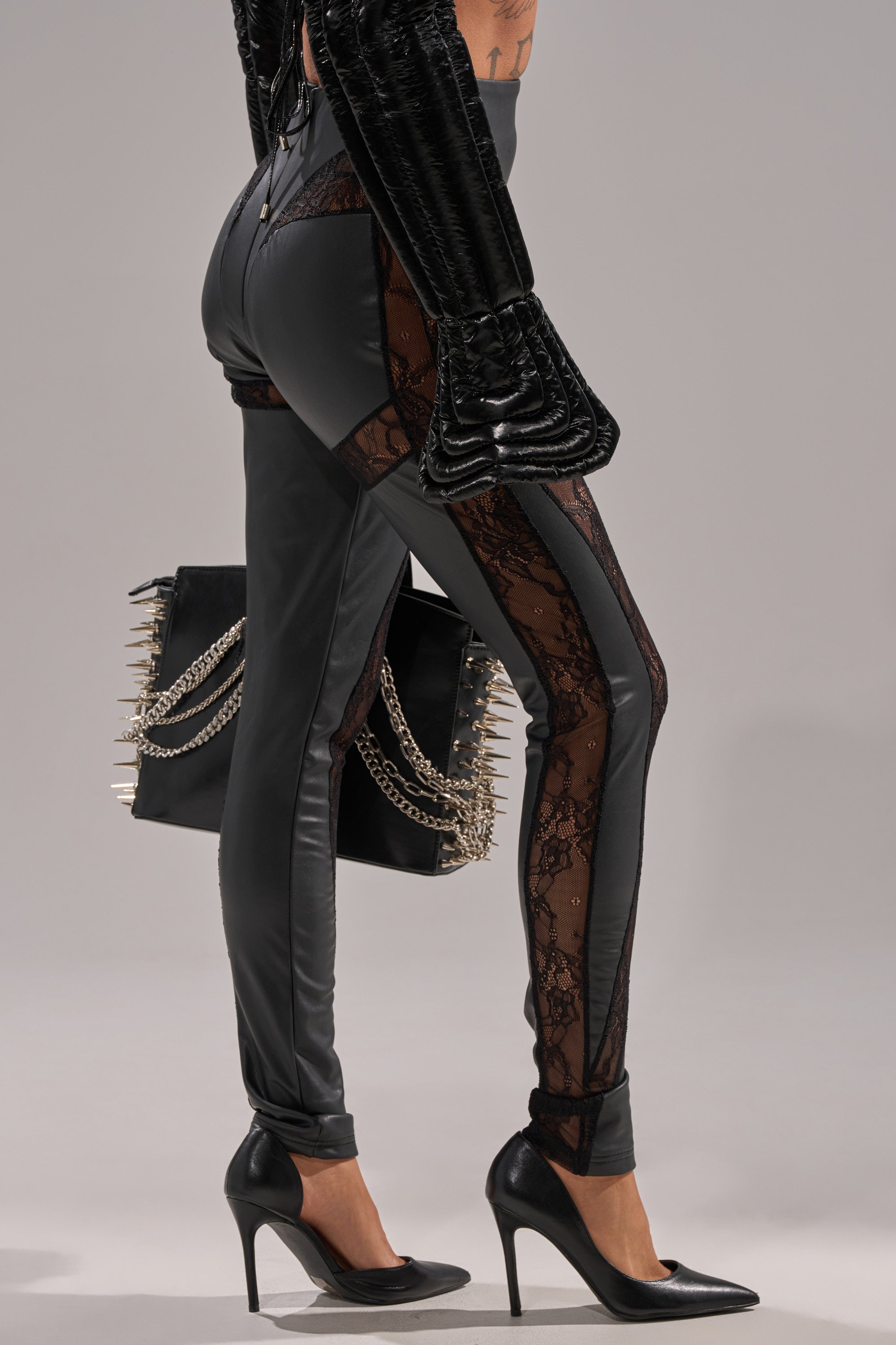 A person wearing LACE DETAIL PANELED FAUX LEATHER LEGGINGS IN BLACK, black high heels, a textured black jacket, and holding a spiked chain-strap black handbag poses against a plain background.