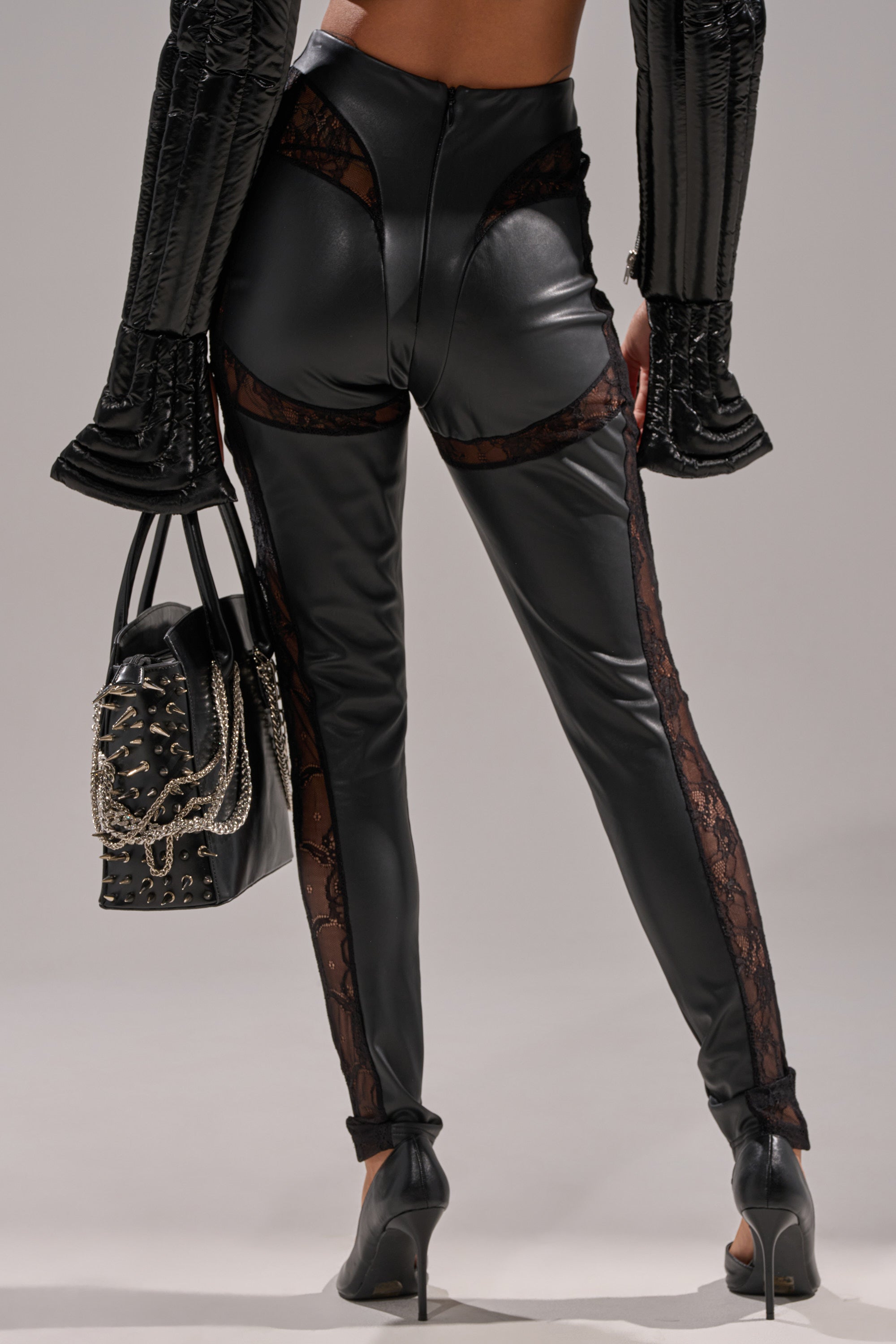 Wearing the LACE DETAIL PANELED FAUX LEATHER LEGGINGS IN BLACK, a person in heels holds a black chain-detailed handbag. The photo, taken from behind, emphasizes the leggings' unique lace-accented back design.