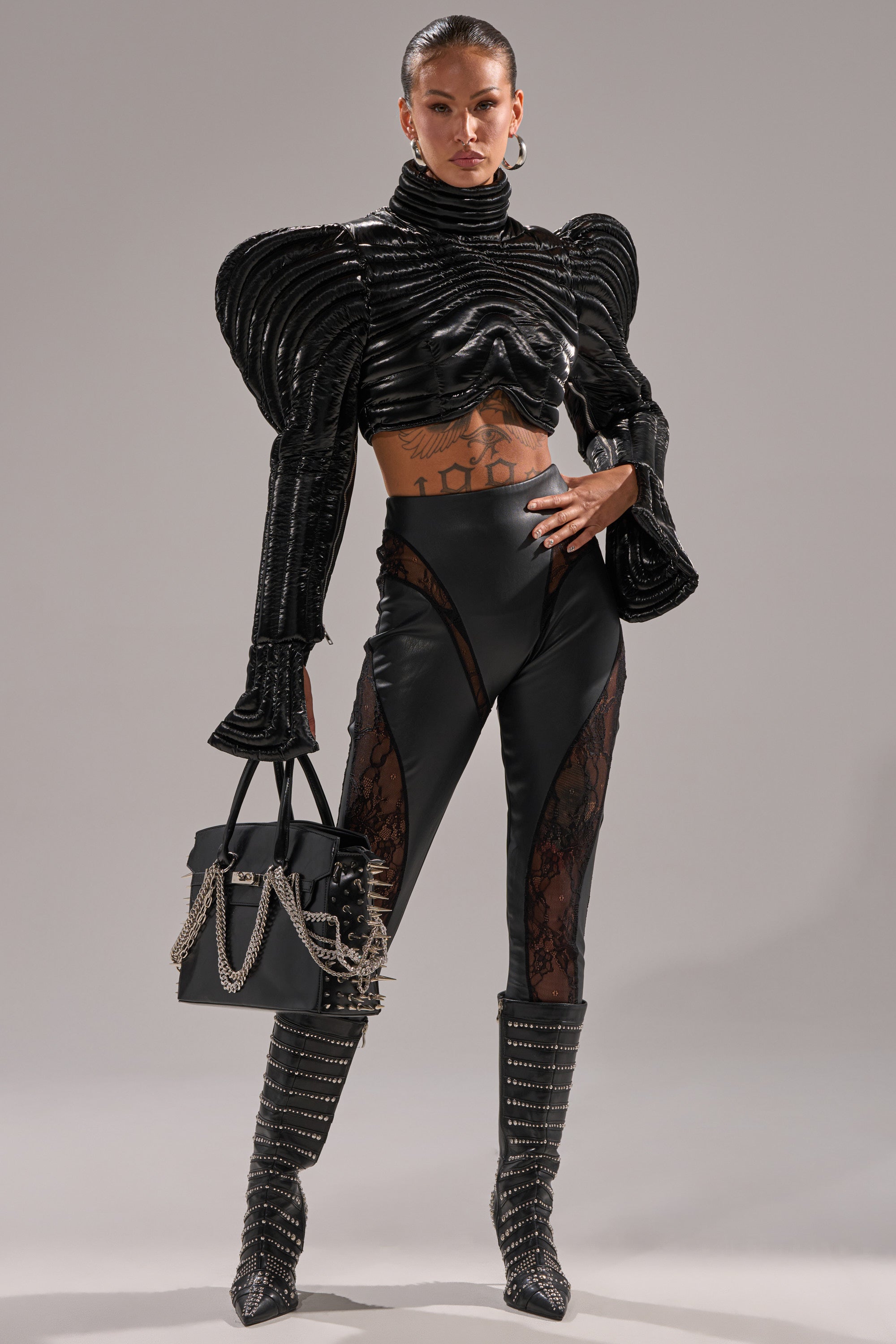 A woman poses in LACE DETAIL PANELED FAUX LEATHER LEGGINGS IN BLACK, styled with a bold black top, studded boots, and a black chain-detailed handbag, showcasing her tattoos.