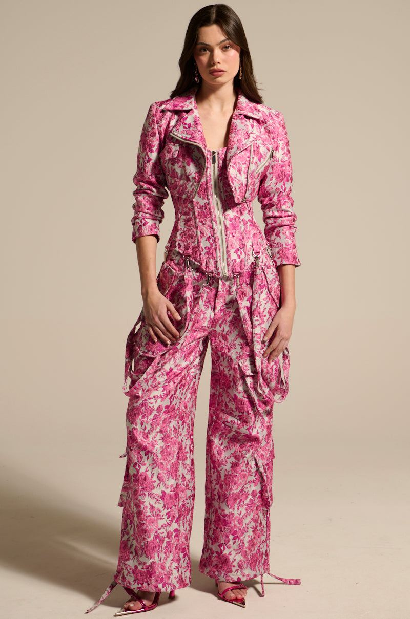 A person models vibrant pink jumpsuit-style MILEY JACQUARD CARGO PANTS with zippered details, a fitted bodice, and high-rise wide-leg pants featuring floral jacquard fabric. Pink shoes complete the look against a plain, neutral background. 0
