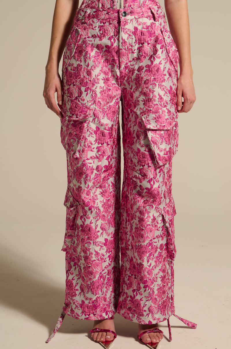 Someone is donning high-rise Miley Jacquard Cargo Pants in a pink and white floral pattern, featuring multiple pockets and tie details at the ankles. They pair them with red strappy sandals against a neutral beige background. 2