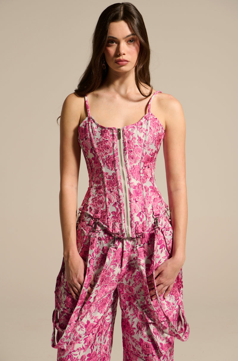 A person with long brown hair wears a pink Miley Jacquard Corset top and matching wide-leg pants, featuring an intricate pattern, zipper detail, and floral jacquard fabric that fits perfectly against the plain beige background.