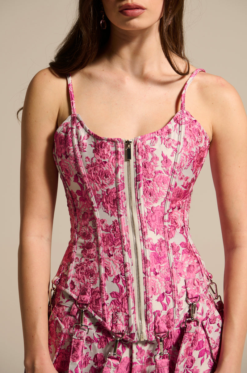 A woman in the Miley Jacquard Corset, featuring a pink floral jacquard design with a curve-hugging fit, thin straps, and front zipper. Her matching shorts enhance the rose pattern while her long dark hair cascades over her shoulders against a beige background.