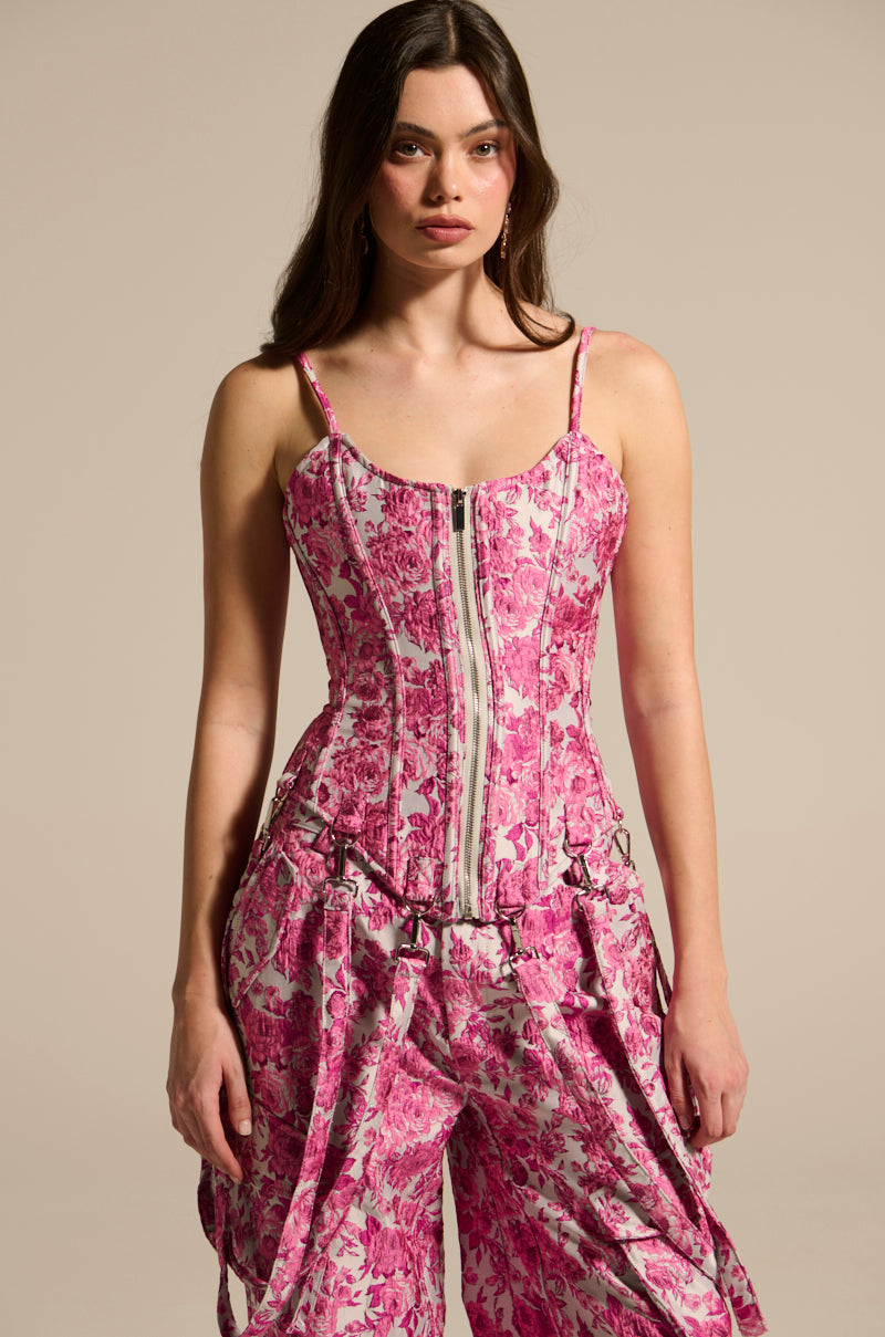 A woman models the Miley Jacquard Corset, featuring a pink and white floral jacquard design with a front zipper. The curve-hugging fit complements loose, draped pants as her long dark hair cascades against the neutral backdrop.