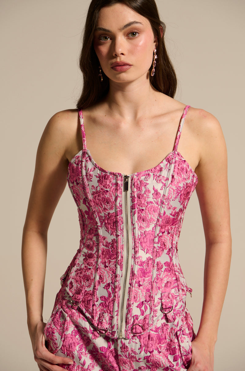 A woman with long dark hair models a pink MILEY JACQUARD CORSET featuring zipper details and matching pants, showcasing the curve-hugging floral jacquard fabric against a neutral background as she stands with her hands by her sides, gazing at the camera.