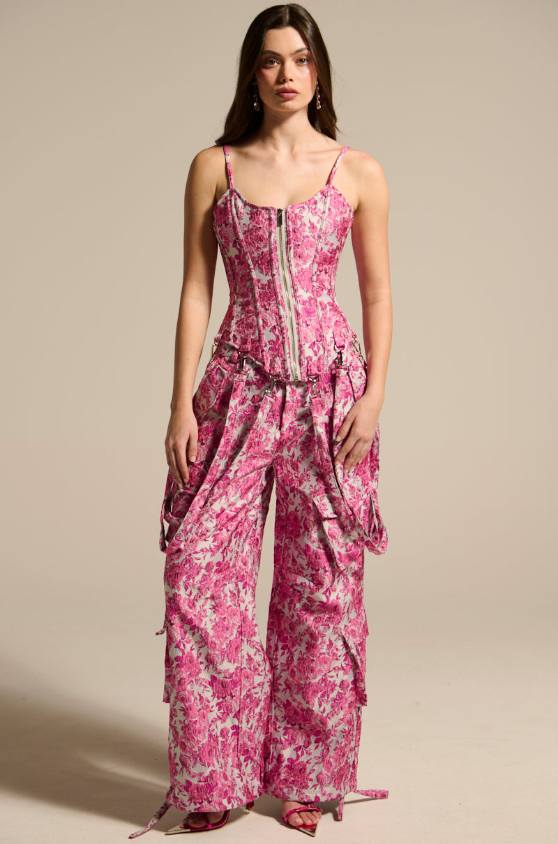 Wearing the Miley Jacquard Corset in a pink floral jacquard outfit, a woman poses gracefully. Her curve-hugging corset is complemented by flowy ruffled pants and cascading hair against a neutral background.