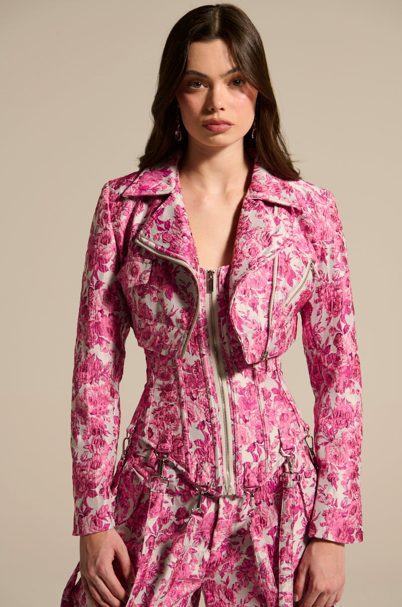 A woman flaunts a curve-hugging fit in the MILEY JACQUARD CORSET, a stylish pink and white floral jacquard jacket with a matching skirt. The zip design and structured form complement her long brown hair against a neutral background.
