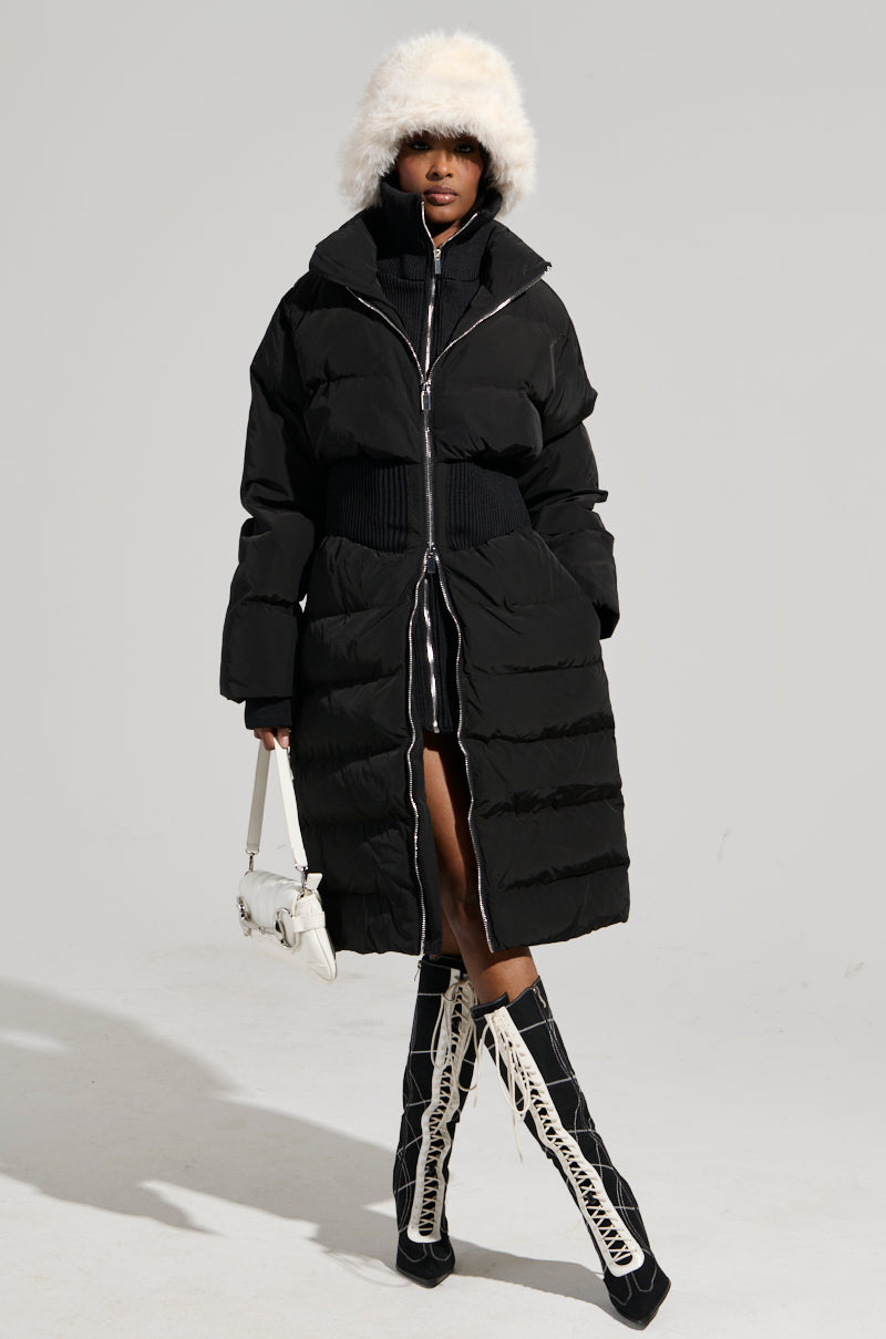 A person wearing the STASSI FITTED PUFFER by AKIRA Label, featuring a metallic front zipper closure, a white furry hat, and knee-high black and white lace-up boots stands against a plain background, holding a small white handbag.
