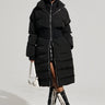 A person wearing the STASSI FITTED PUFFER by AKIRA Label, featuring a metallic front zipper closure, a white furry hat, and knee-high black and white lace-up boots stands against a plain background, holding a small white handbag.