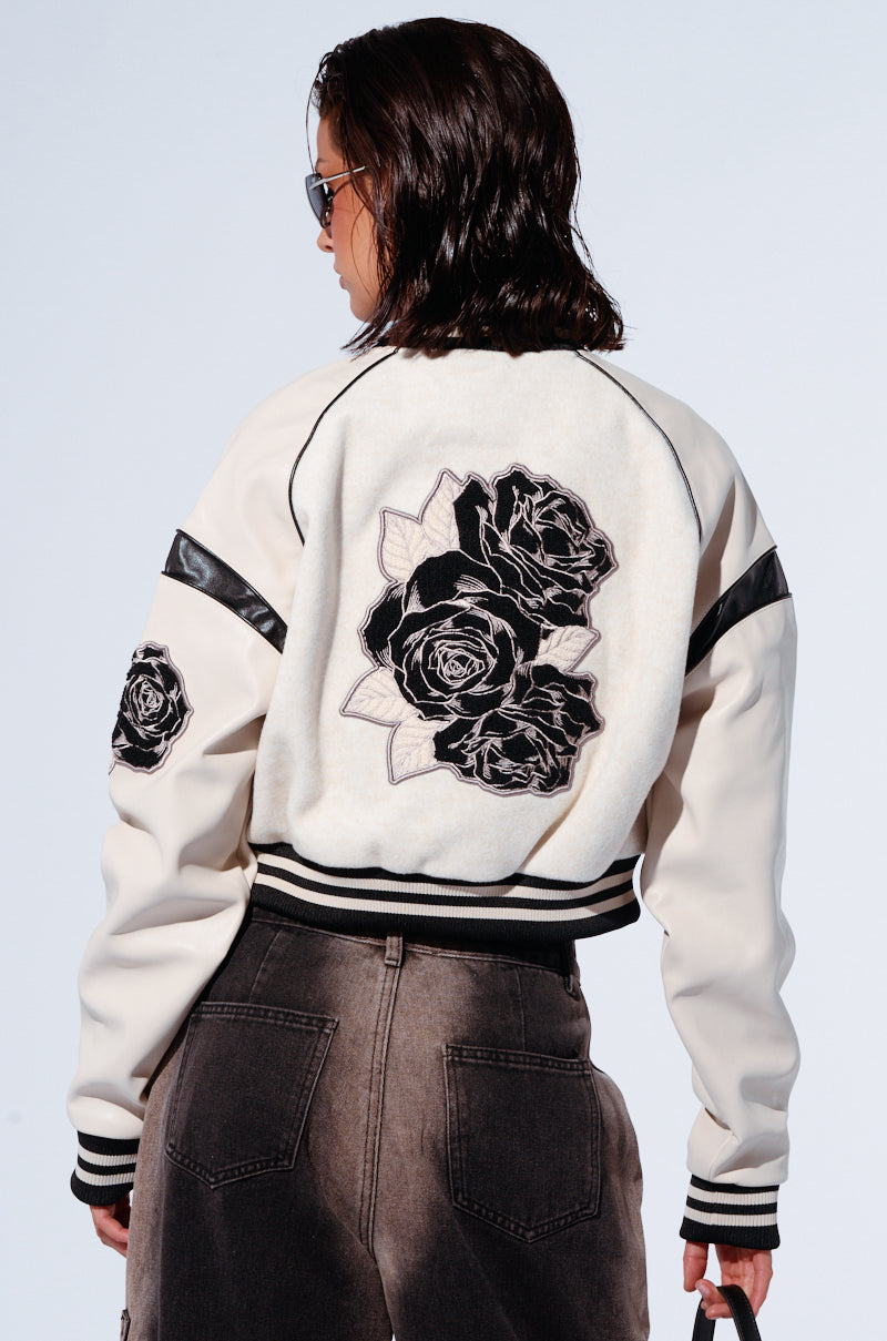 A person with shoulder-length dark hair, wearing sunglasses, is seen from the back. They are dressed in the EVERY ROSE EMBROIDERED BOMBER jacket, which features large black rose designs on the back and a left faux leather sleeve, paired with dark gray jeans. The person carries a small black item in their left hand.