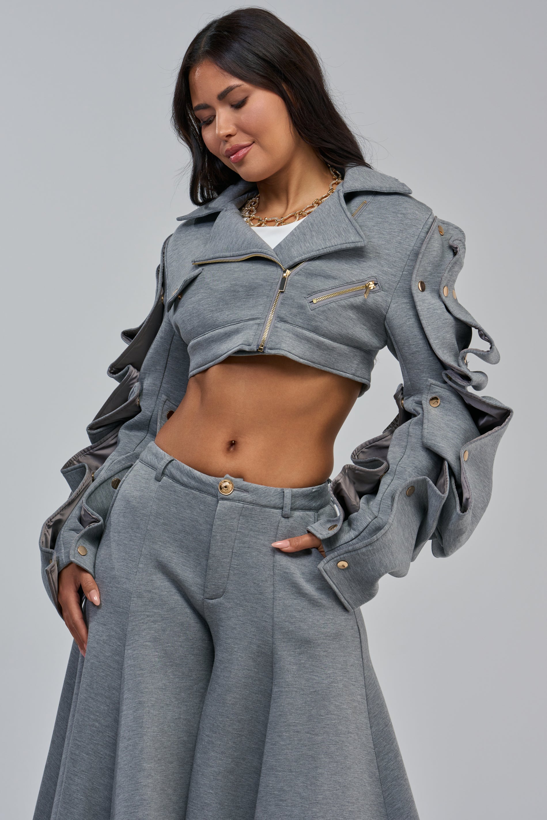 A woman wears the SNAP OUT OF IT ULTRA CROP MOTO IN SCUBA—a stylish grey cropped moto jacket with ruched sleeves—paired with matching high-waisted pants, standing against a plain background and looking down with a slight smile.