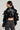 A woman stands with her back to the camera, wearing the SNAP OUT OF IT ULTRA CROP MOTO faux leather jacket that features ruffled, voluminous sleeves. She pairs it with high-waisted, dark gray jeans and large gold hoop earrings. Her hair is styled in a sleek low ponytail.