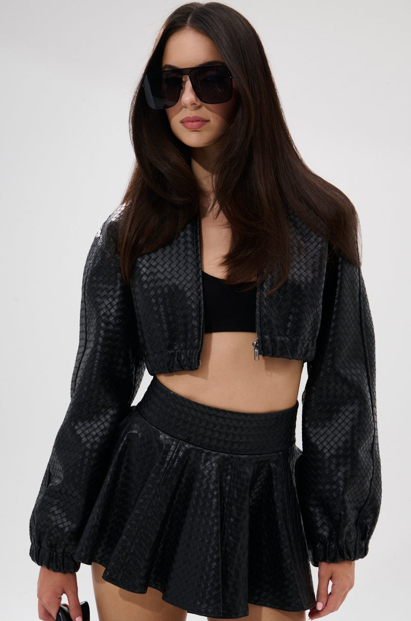 A woman with long brown hair stands against a plain light background, wearing oversized black sunglasses, a black crop top, and the KYLIE HOODED BOMBER paired with a matching high-waisted mini skirt. 1