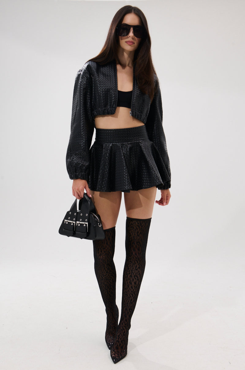 A woman wearing the AKIRA Label KYLIE HOODED BOMBER, a matching mini skirt, black crop top, patterned over-the-knee boots, sunglasses, and a black handbag stands against a plain white background. 9