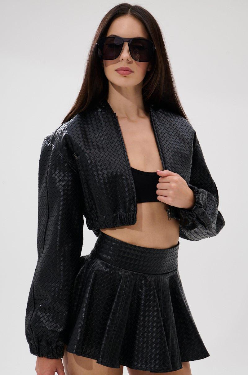 A woman with long dark hair models oversized sunglasses, a KYLIE HOODED BOMBER black textured cropped jacket, a black crop top, and a matching high-waisted pleated mini skirt against a plain light background. 3