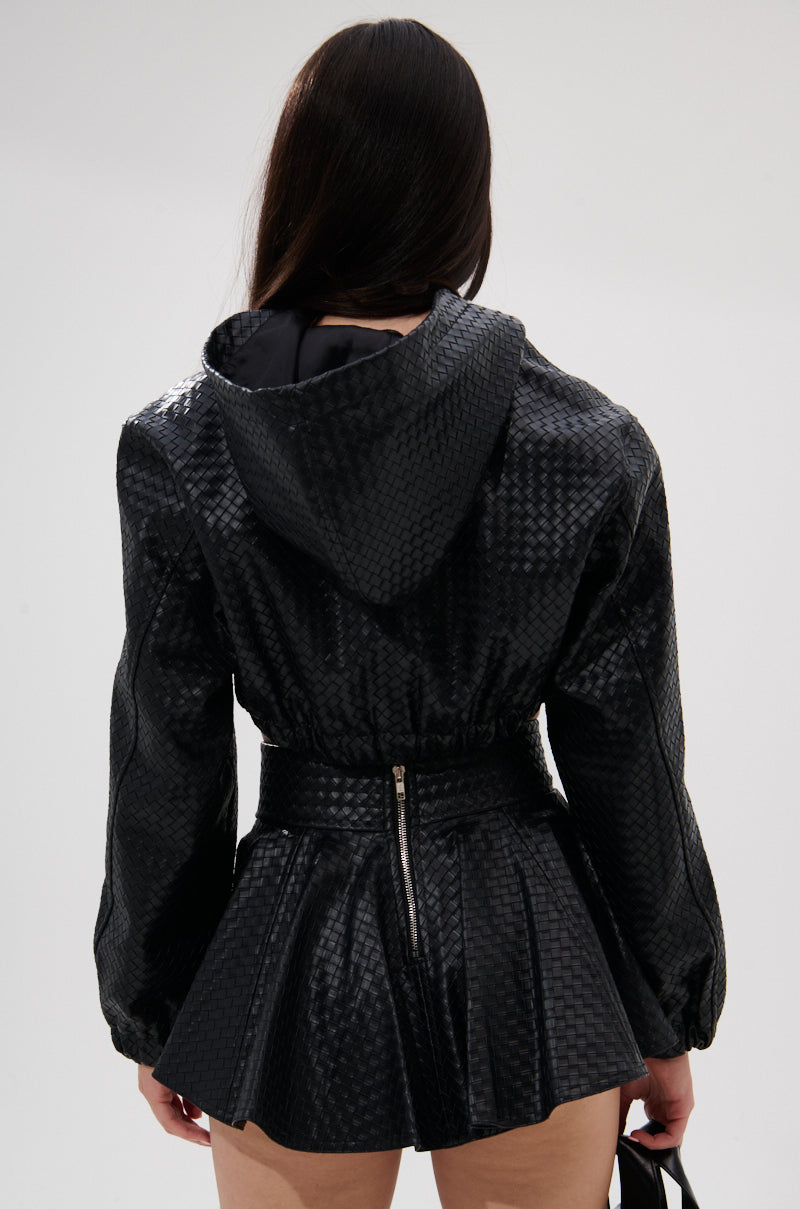A woman with long dark hair stands facing away, wearing the KYLIE HOODED BOMBER—a textured black jacket with hood—paired with a matching pleated skirt featuring a visible back zipper, set against a plain light background. 7