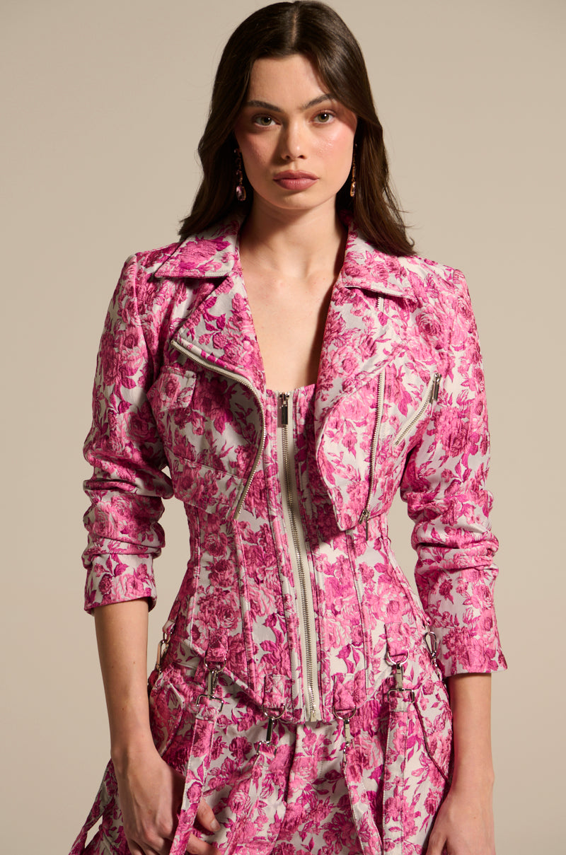 Dressed in the MILEY JACQUARD CROPPED MOTO, featuring a fitted pink and white floral pattern, wide collar, zipper detail, and satin lining. This elegant jacket pairs seamlessly with a matching skirt over a neutral beige background.
