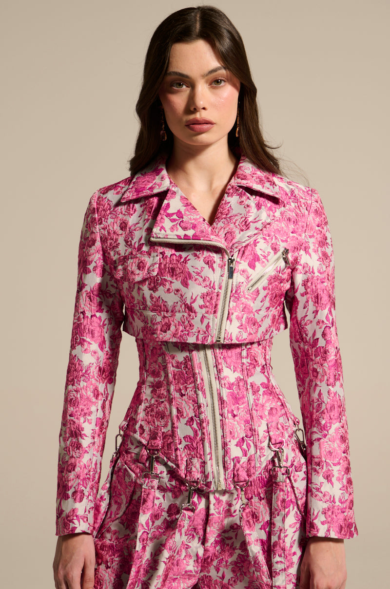 A woman poses in the MILEY JACQUARD CROPPED MOTO, a pink and white floral jacket with visible zippers, set against a neutral backdrop. Her long dark hair cascades down, complemented by subtle makeup.