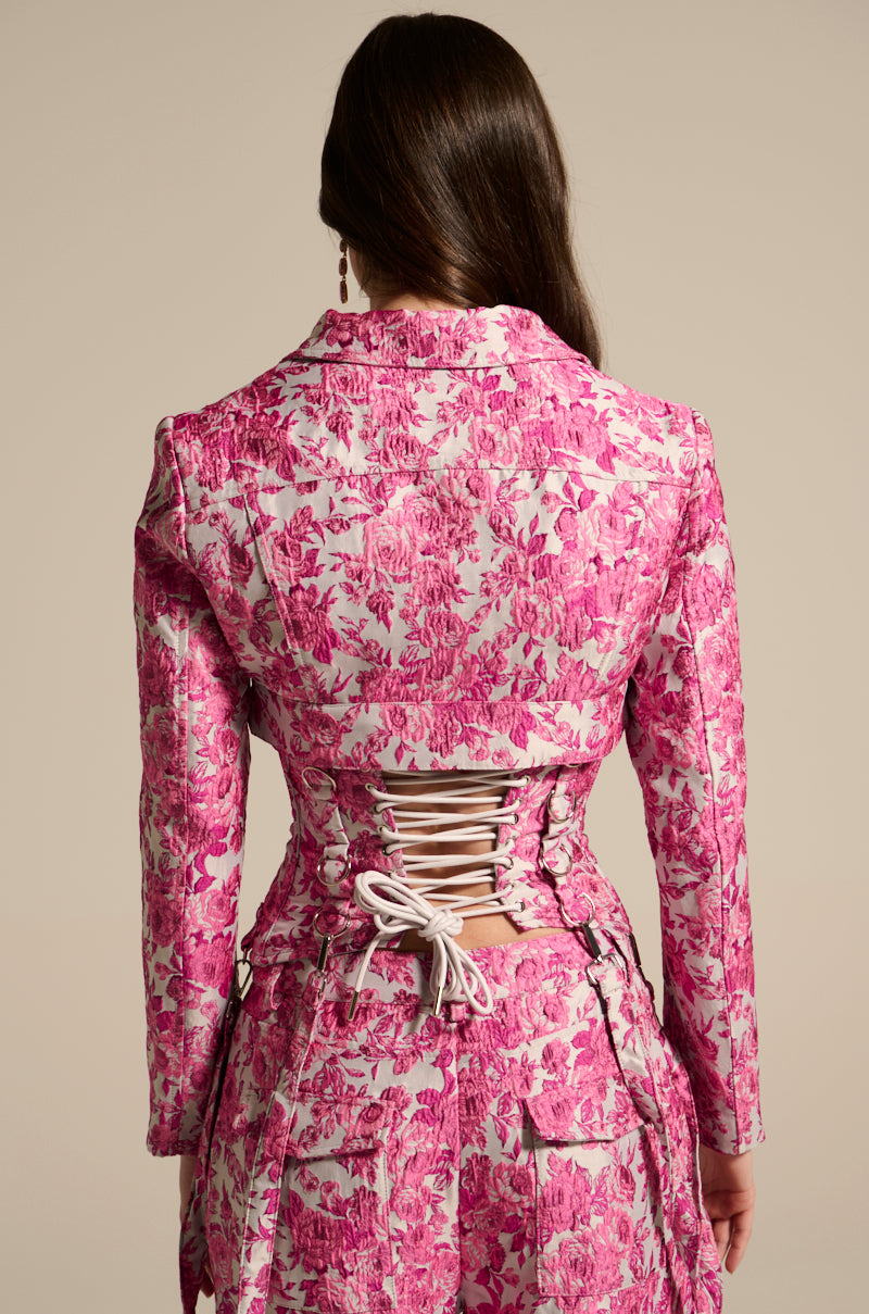 A person with long hair faces away, wearing the MILEY JACQUARD CROPPED MOTO, a pink floral jacket with a satin lining and lace-up back detail. The plain background accentuates the vibrant and intricate design of this unique outfit.