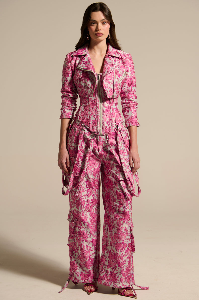 In a pink floral jumpsuit with layered details and a zippered front, a person stands confidently, their long hair styled loose. They wear the MILEY JACQUARD CROPPED MOTO jacket over it, creating an edgy contrast against the plain backdrop.