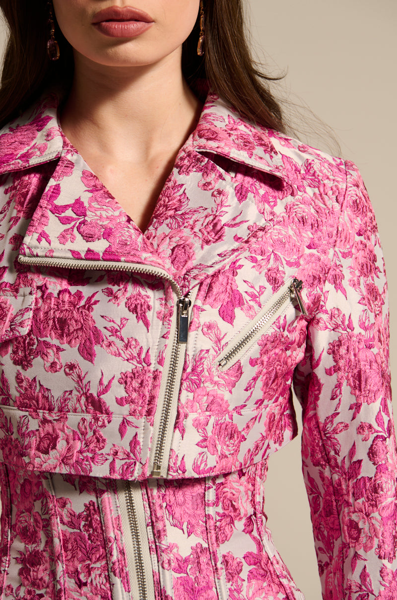 The image shows a woman in the MILEY JACQUARD CROPPED MOTO, a chic pink and white floral jacket with a prominent zipper and notch collar. The fabric features detailed pink flowers. Only the lower half of her face and upper torso are visible, highlighting this satin-lined piece's elegance.