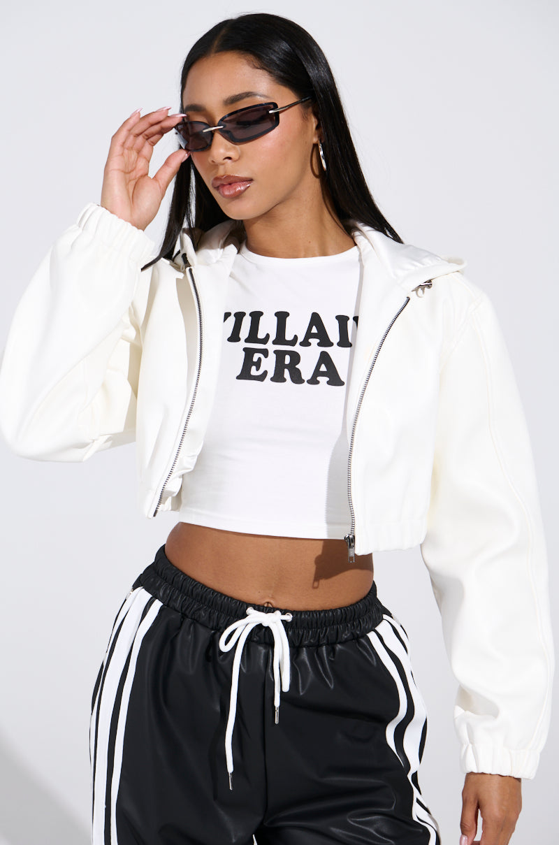 A woman in sunglasses, a "Villain Era" shirt, and the white DEX Hooded Bomber jacket poses against a plain background, adjusting her sunglasses with one hand while wearing black track pants with white stripes.