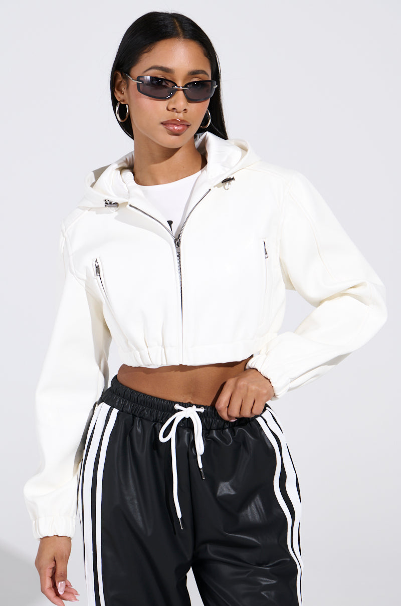 Wearing a DEX Hooded Bomber in white and black track pants with white stripes, a person poses confidently. They have long dark hair, oval sunglasses, and large hoop earrings against a plain light gray background.