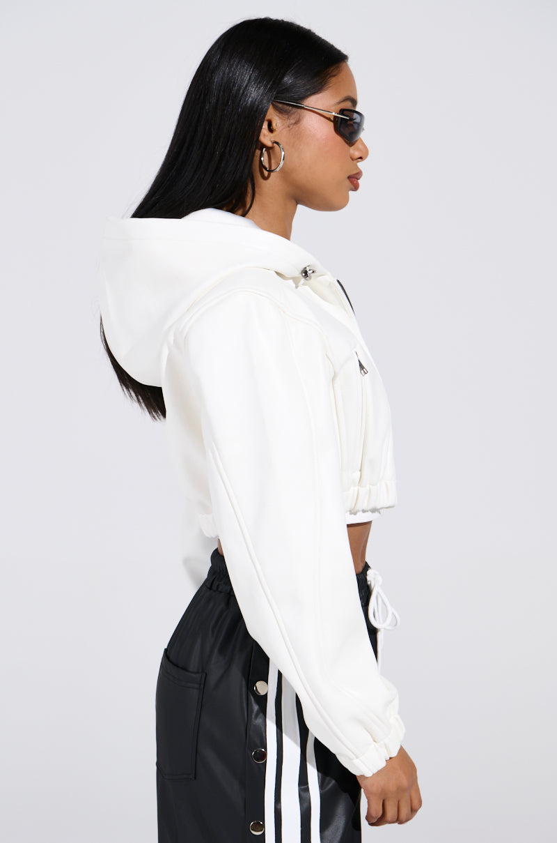 A woman in a DEX HOODED BOMBER IN WHITE and black sunglasses is turned to the side, stylishly highlighting her silhouette. She wears black pants with white stripes, hoop earrings, and has long black hair against a plain, light-colored background.