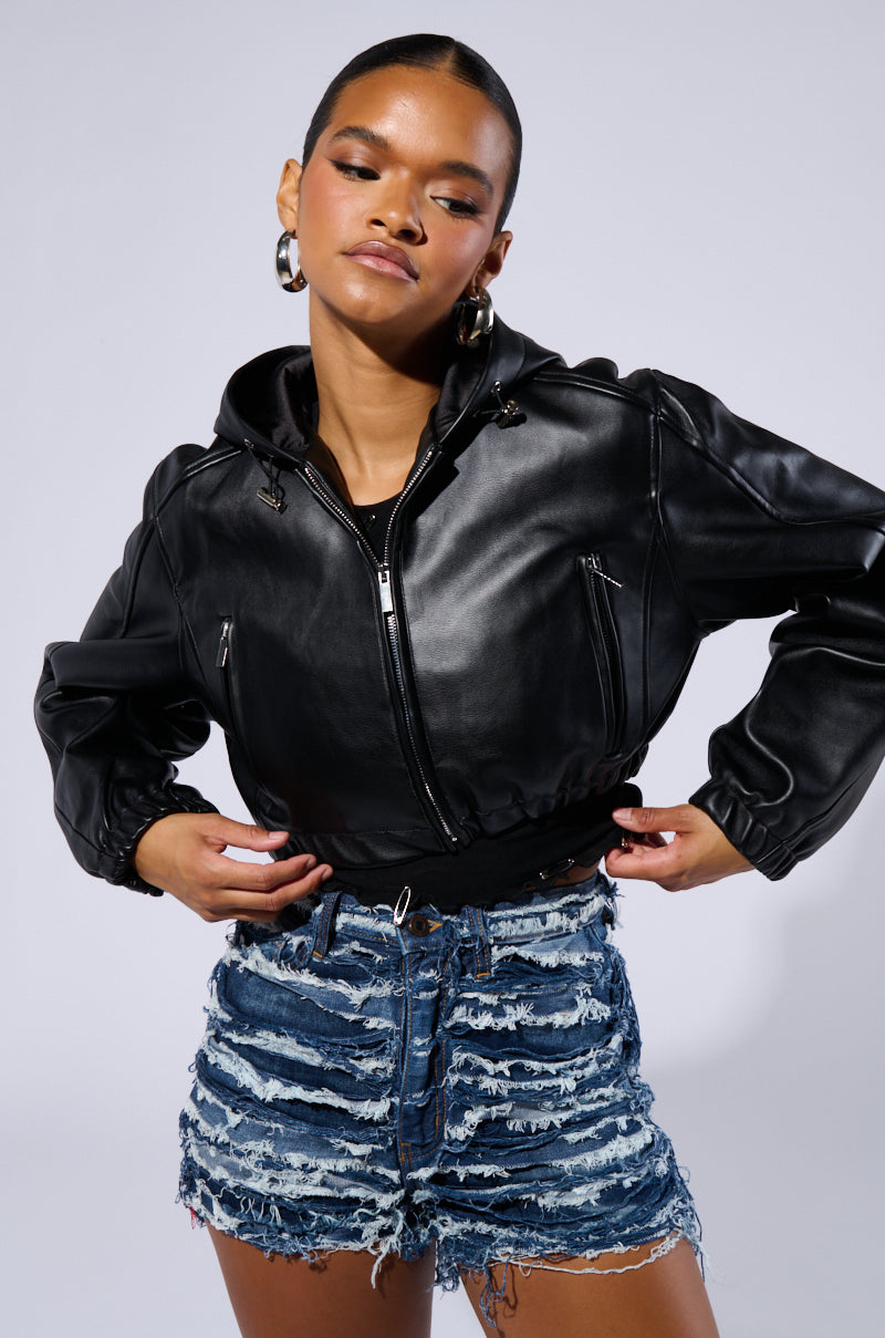 Person wearing the DEX HOODED BOMBER, featuring zipped pockets, paired with high-waisted, heavily distressed denim shorts. The individual stands posed with hands on hips, gazing slightly downward in a plain studio setting.