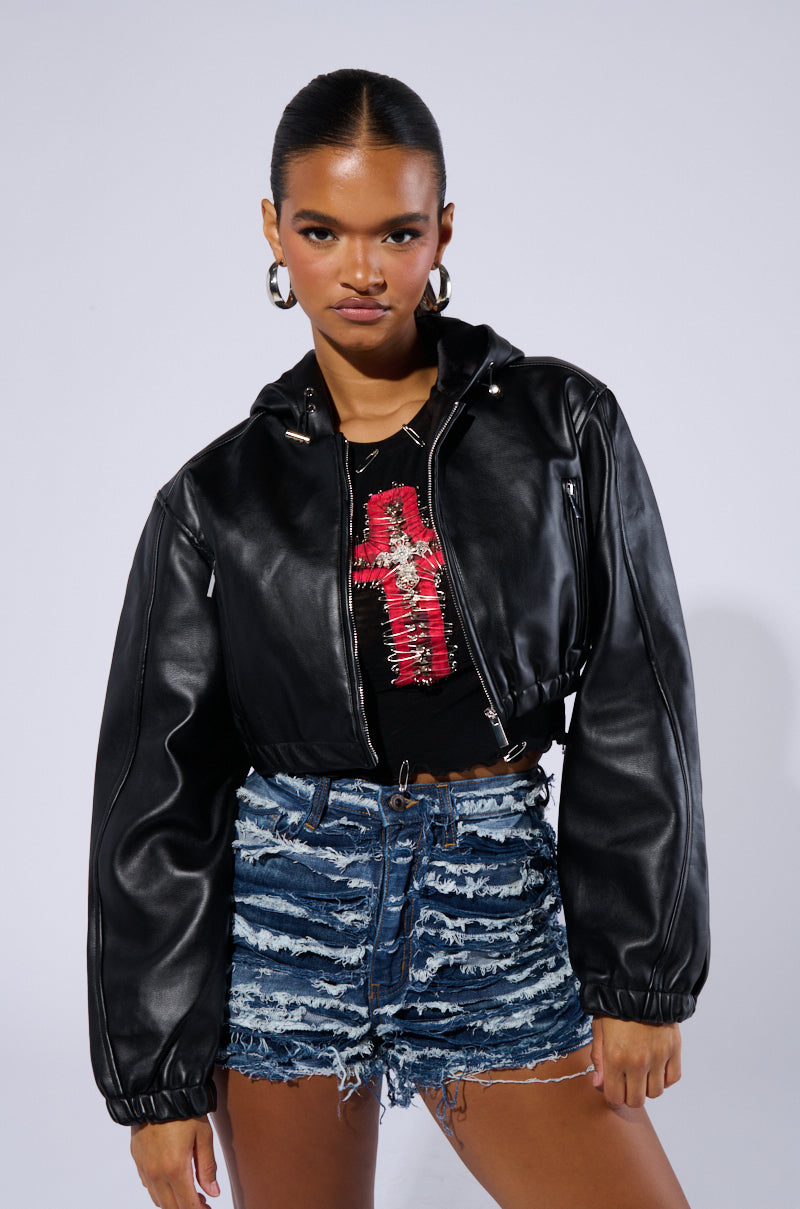 A person with dark hair pulled back, wearing hoop earrings, a DEX HOODED BOMBER jacket over a black top with a red design, and distressed denim shorts. They are standing against a neutral background with a confident expression.