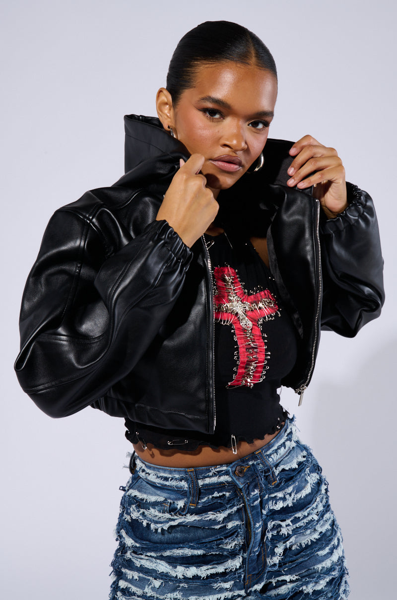 A person with neat hair wears the DEX HOODED BOMBER, a black faux leather jacket with a high collar, over a black crop top featuring a red cross design. They pair it with blue distressed denim jeans and are adjusting the collar while facing the camera with a confident expression.