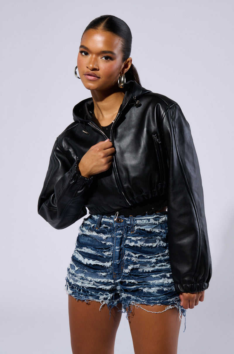 A woman with dark hair tied back, wearing large hoop earrings, the DEX HOODED BOMBER jacket, and distressed denim shorts stands against a plain background. She holds part of her jacket with one hand, gazing confidently at the camera.