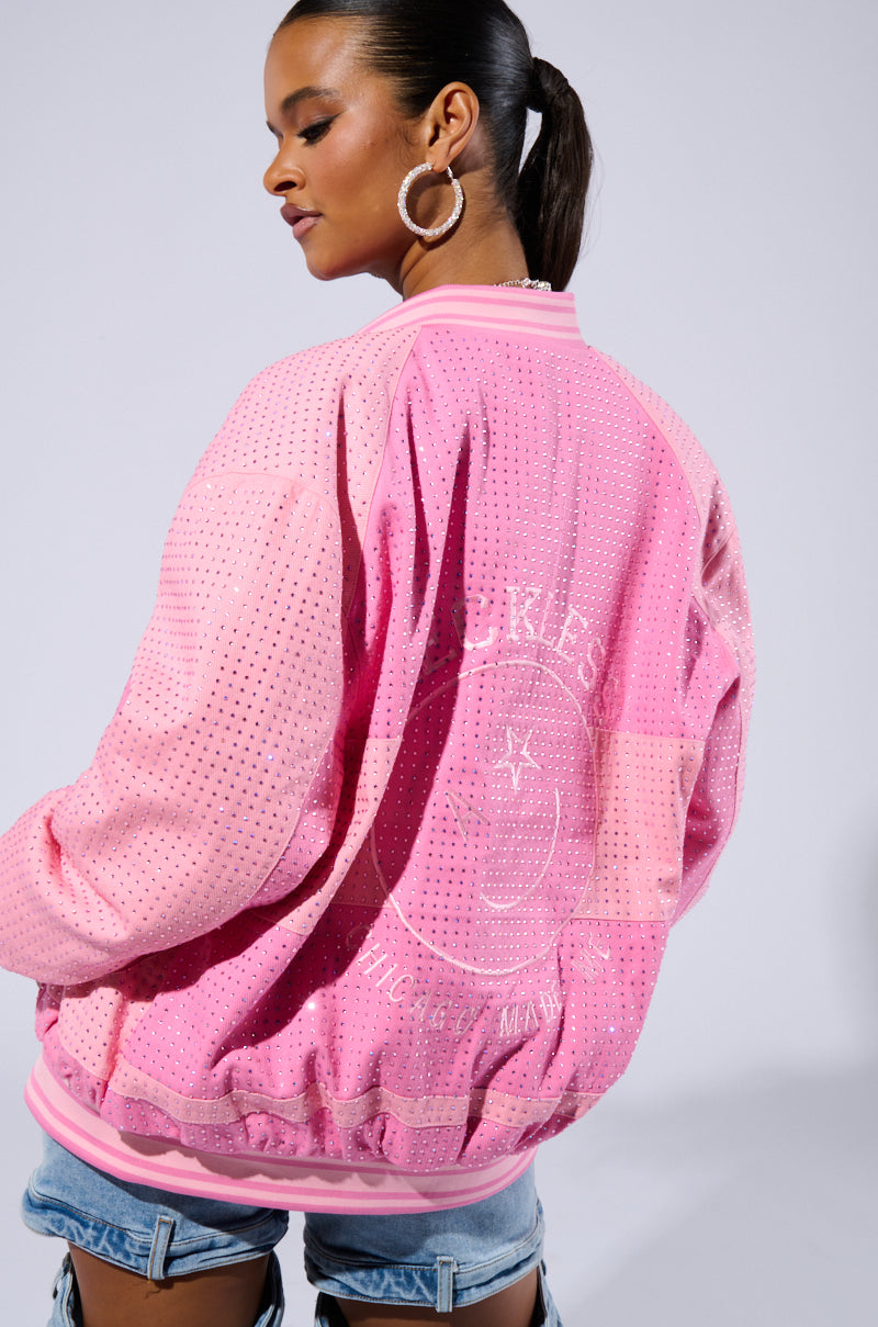 A woman stands facing slightly to the side, wearing a vibrant pink, studded "Chicago Made Embroidered Bomber" with a circular pattern and text on the back. Her hair is tied back in a ponytail, complemented by large hoop earrings and denim shorts. The background is plain and well-lit.