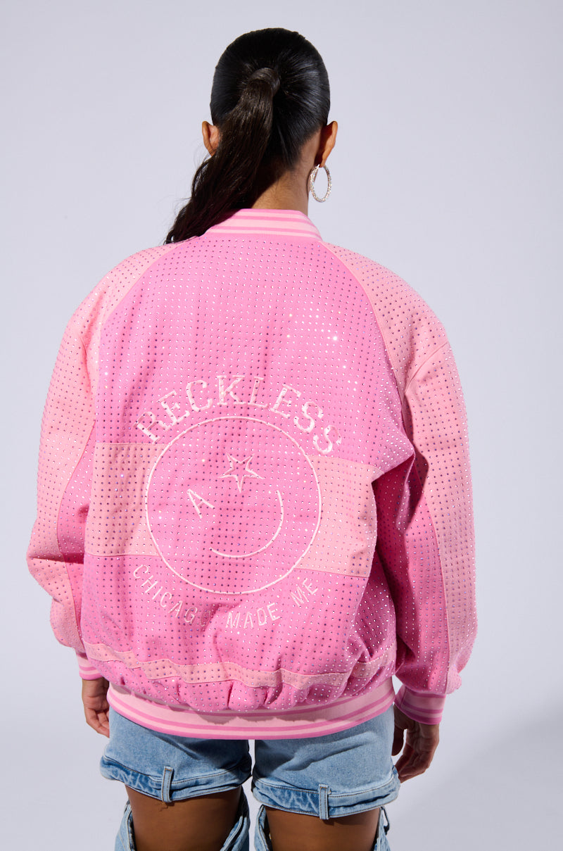 A person with long, dark hair is seen from the back, wearing a pink, perforated "CHICAGO MADE EMBROIDERED BOMBER" jacket with a quilted satin inner lining. The jacket features the word "RECKLESS" and a smiley face design on the back. The person is also sporting hoop earrings and light-washed denim jeans.