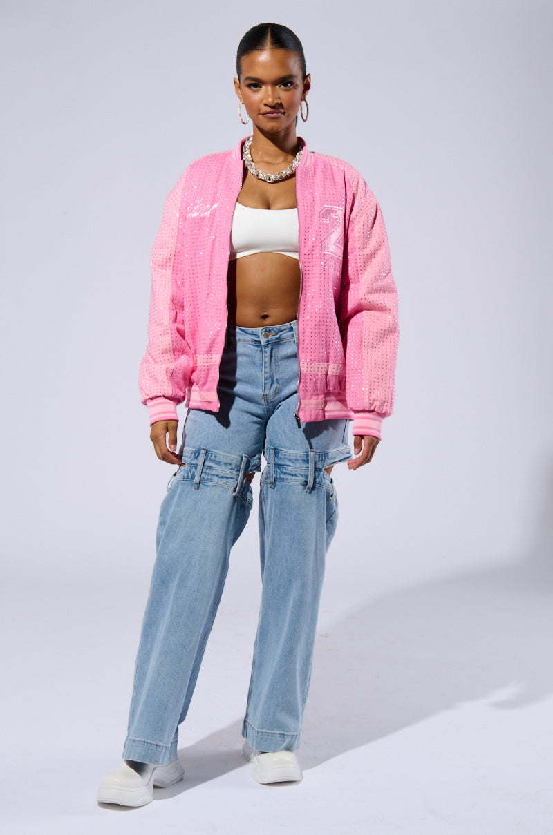 A person stands confidently, showcasing the CHICAGO MADE EMBROIDERED BOMBER with its quilted satin inner lining. They pair it with a white crop top and light blue baggy jeans adorned with zipper details. Complementing the outfit, they wear white shoes and a stylish necklace, while their hair is pulled back tightly. The plain gray background adds to the modern aesthetic.