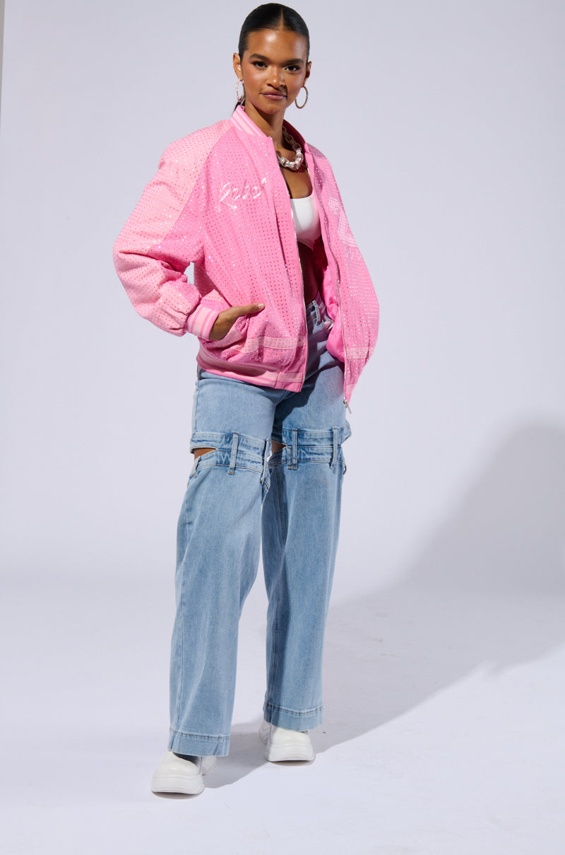 A person stands confidently wearing the Chicago Made Embroidered Bomber in pink over a white top, paired with light blue, wide-leg jeans featuring unique cut-out details. They complete their look with white shoes and hoop earrings, and have their hair pulled back.