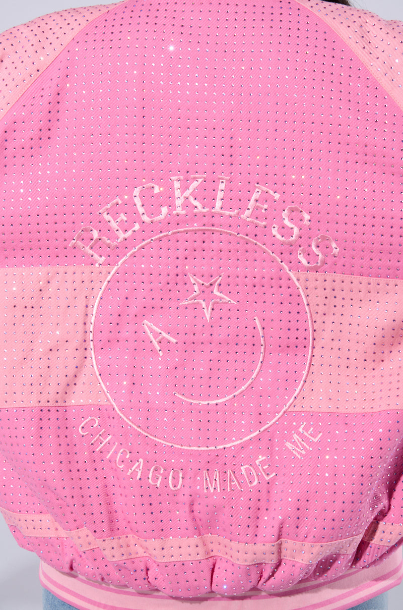 Close-up of the back of the CHICAGO MADE EMBROIDERED BOMBER in pink, showcasing intricate embroidery with "RECKLESS" at the top. Below, an outline of a face features an "A" for one eye, a star for the other, and a smiley mouth. The text "CHICAGO MADE ME" is stitched at the bottom. The jacket is adorned with small, shiny rhinestones throughout and has a quilted design.