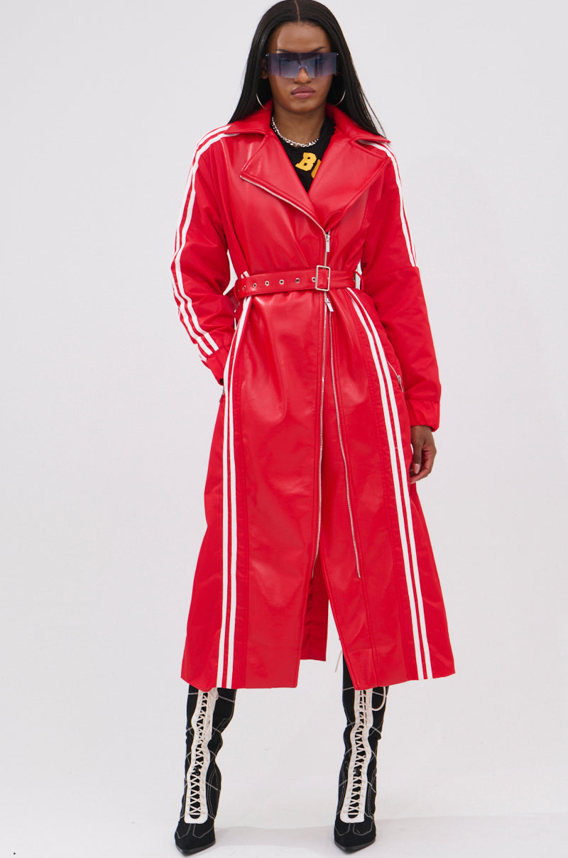 A woman stands confidently against a plain background in the UNAVAILABLE MIXED MEDIA TRENCH, a long red belted coat with white stripes on the sleeves and sides, styled with a black graphic shirt, large sunglasses, and black lace-up heeled boots.