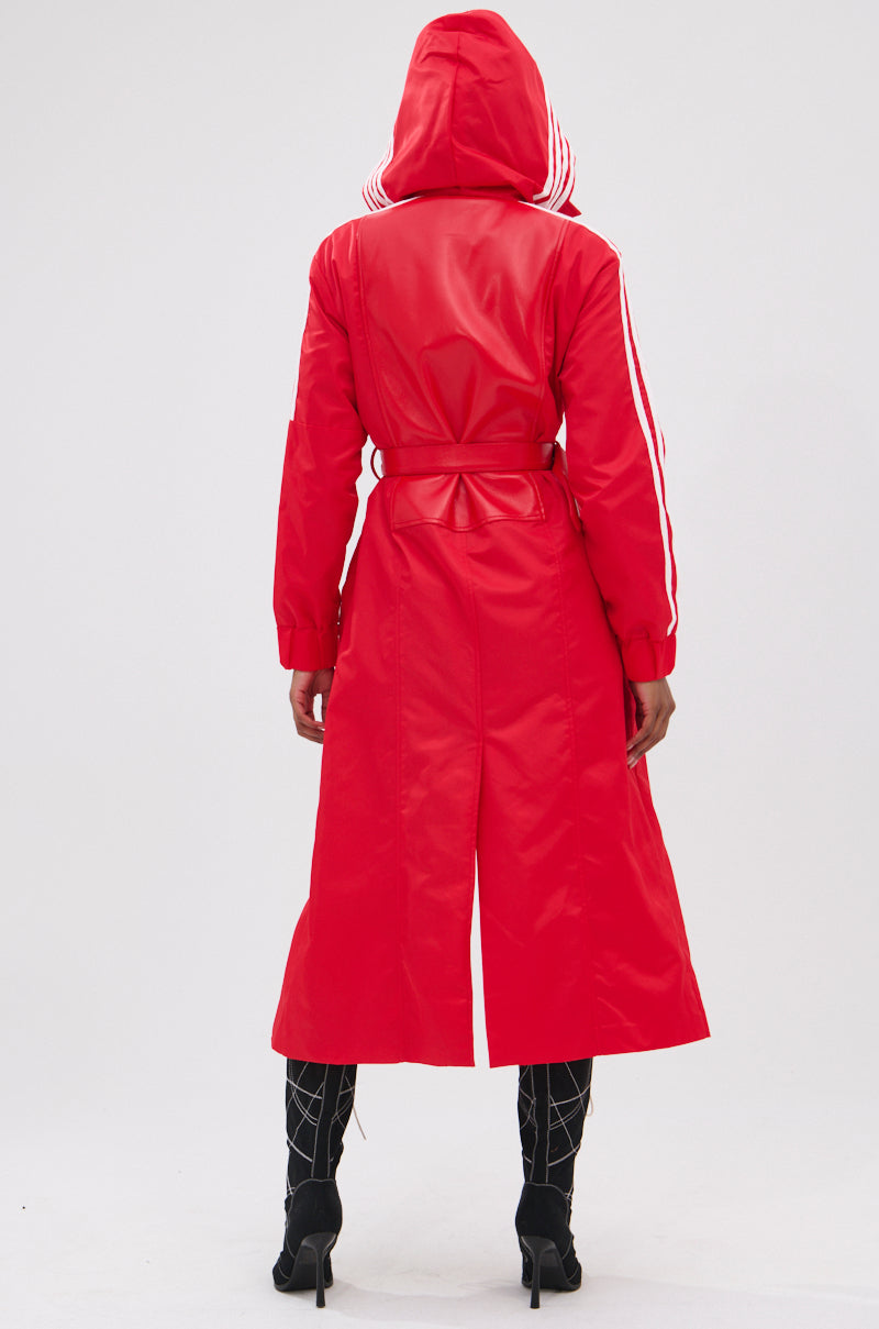 A person stands with their back to the camera, wearing the UNAVAILABLE MIXED MEDIA TRENCH— a long red faux leather hooded coat with white sleeve stripes and a belted waist—paired with black patterned heeled boots against a plain light background.
