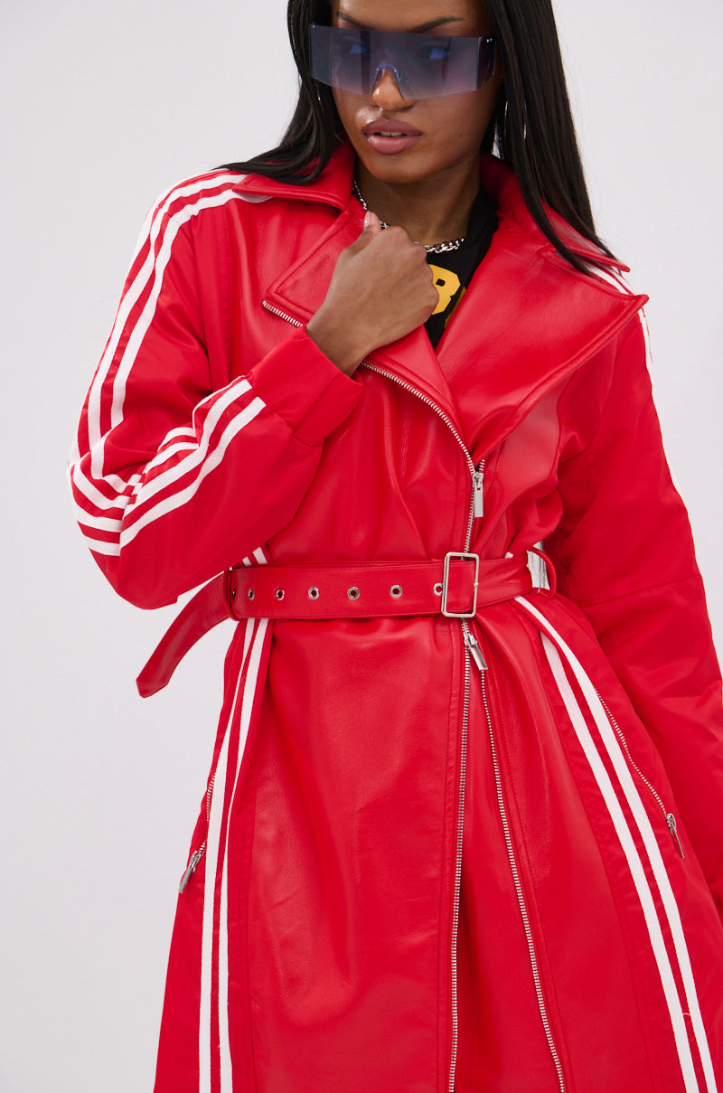 A person with long dark hair and large tinted sunglasses poses with one hand near their collar, wearing the UNAVAILABLE MIXED MEDIA TRENCH—a bright red faux leather belted jacket featuring white sleeve stripes.